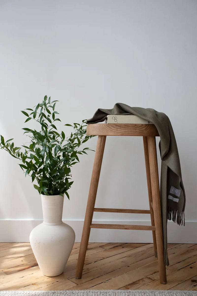 Paris Baby Alpaca Throw in olive green hanging over a wooden stool with a minimalist backdrop