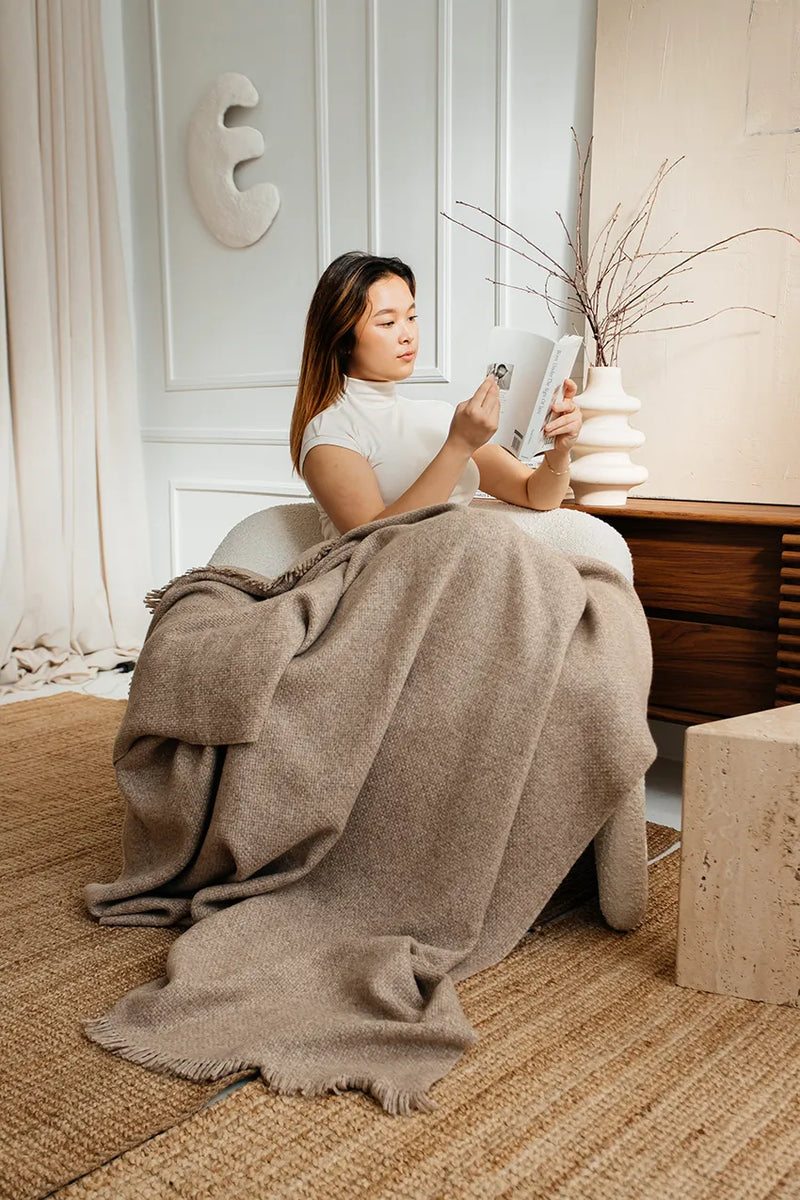 Woman reading a book while wrapped in a beige Upland Textured Baby Alpaca Throw